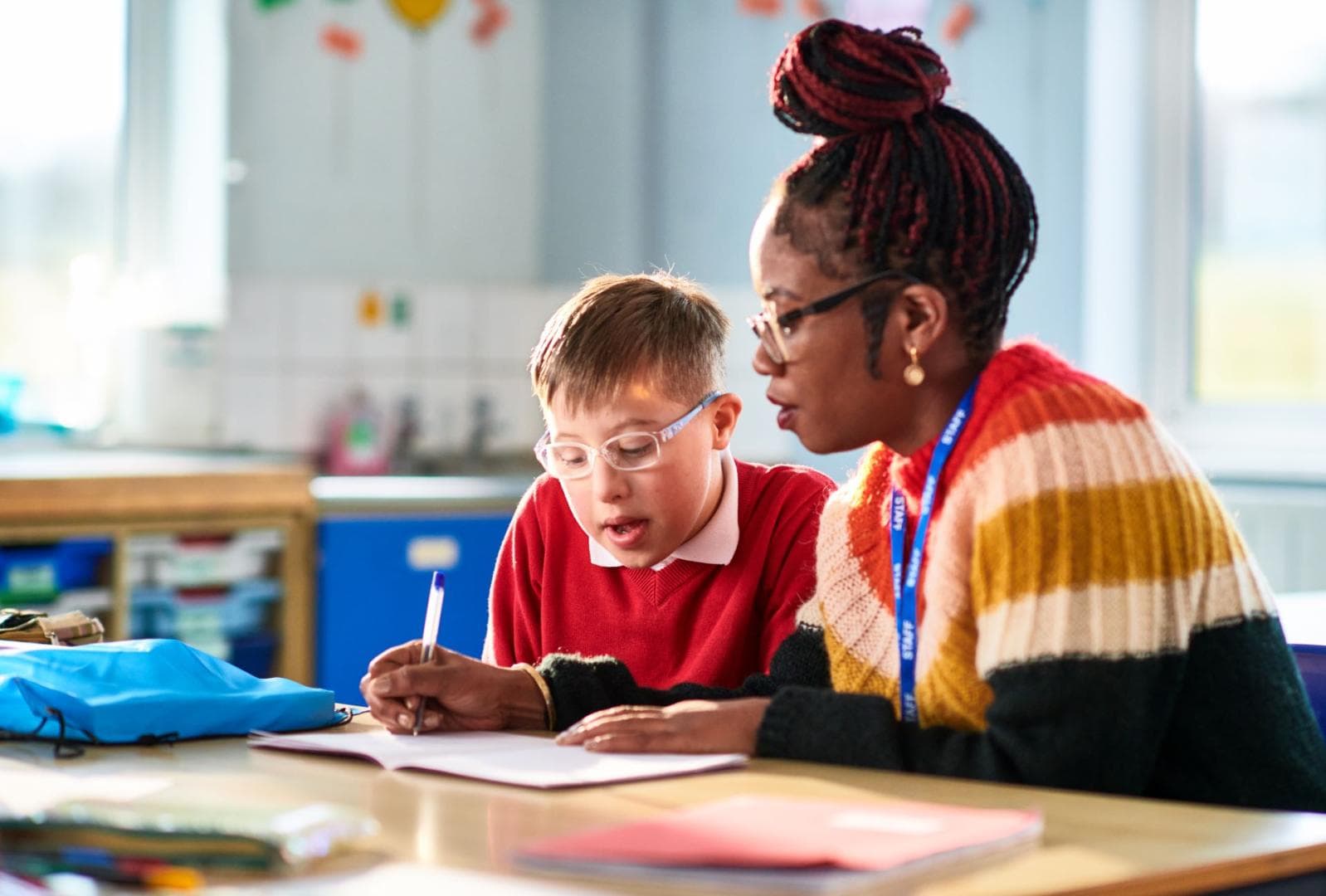 A young boy wearing glasses is sitting next to a teaching assistant in the classroom. They are working on some writing together. 