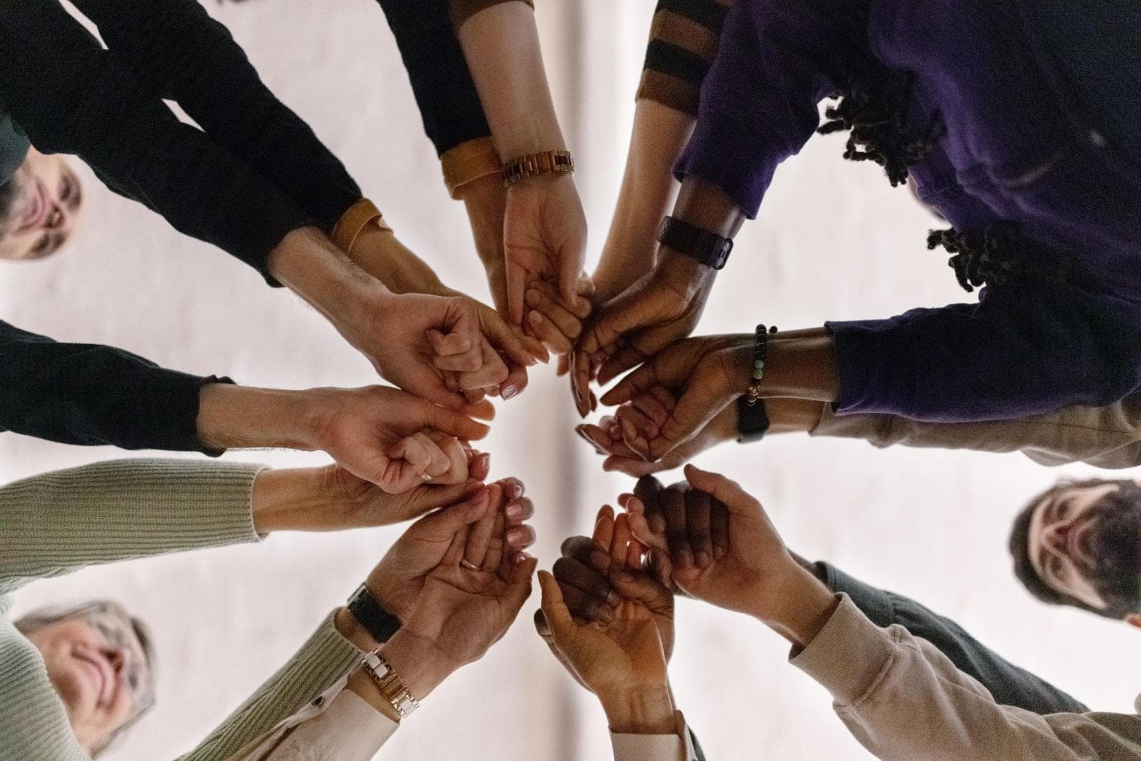 A diverse group of people are standing in a ring. They are all placing their hands in the centre as a sign of unity, 