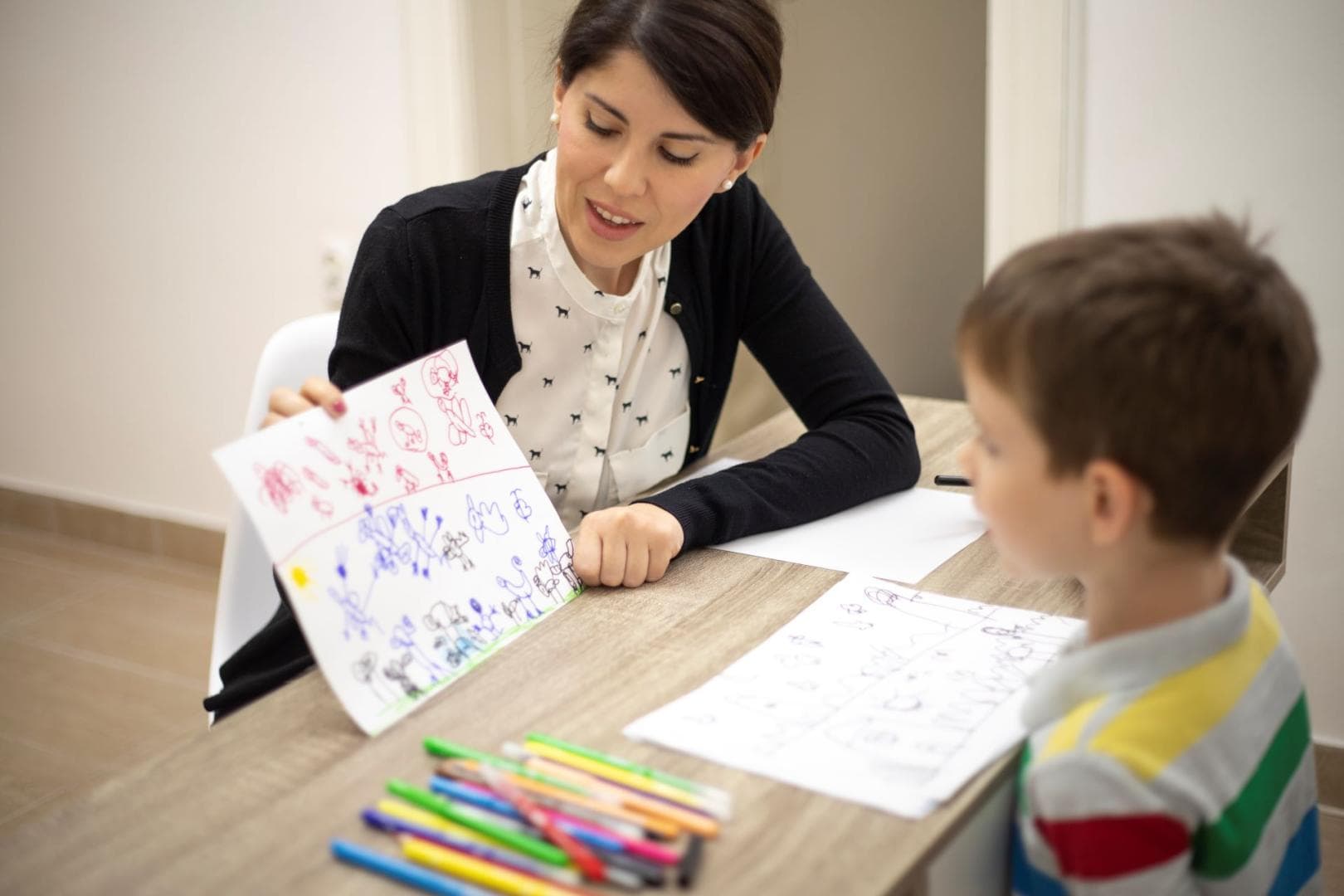 A young boy and a woman are sitting at a desk, opposite to one another. On the desk, there is a pile of coloured crayons. The woman is holding the boys artwork and talking about it with him. 