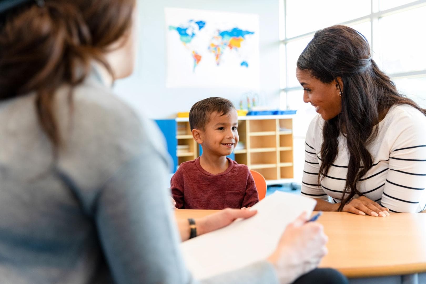 An educational psychologist is sitting at a desk, talking to a young boy. They are both smiling 