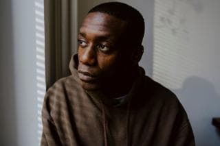 A Black man wearing a brown hooded jumper is staring out the window. He looks sad.