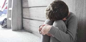 A young boy sat on the floor of his bedroom with his head in his hands feeling sad