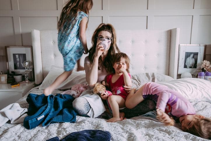 Young mother sipping coffee on bed, surrounded by her three daughters