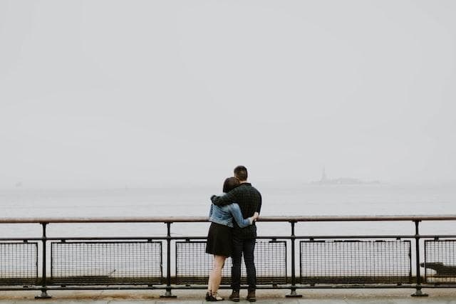 The backs of a man and woman with an arm around each other looking out over a foggy body of water. 