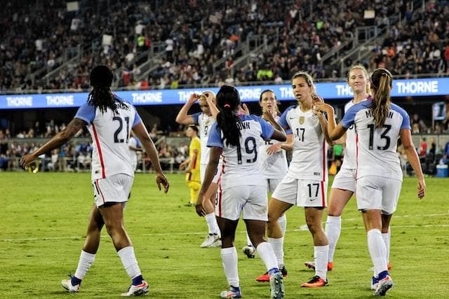 Women's football team on a pitch with a large crowd behind them.