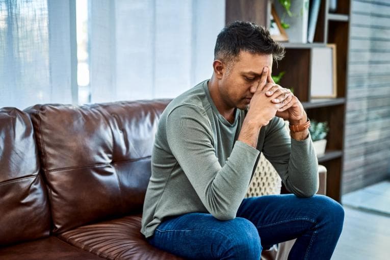 A Indian man wearing blue jeans and a green top is sitting on a sofa. He has his hands clasped together in front of his face and he looks stressed and sad. 