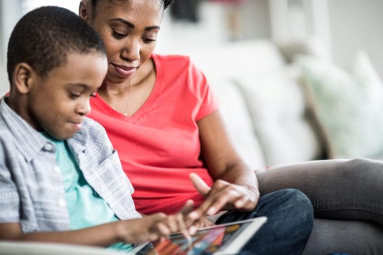 A woman and a boy are sitting on a sofa. They are looking at an iPad together. 