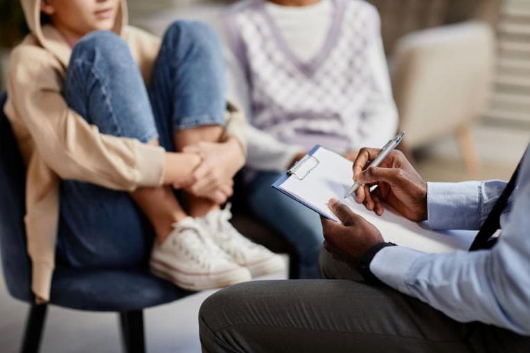 Two teenagers are in a therapy session. One of the teenagers is sitting with her legs tucked up onto the chair. The therapist is taking notes. 