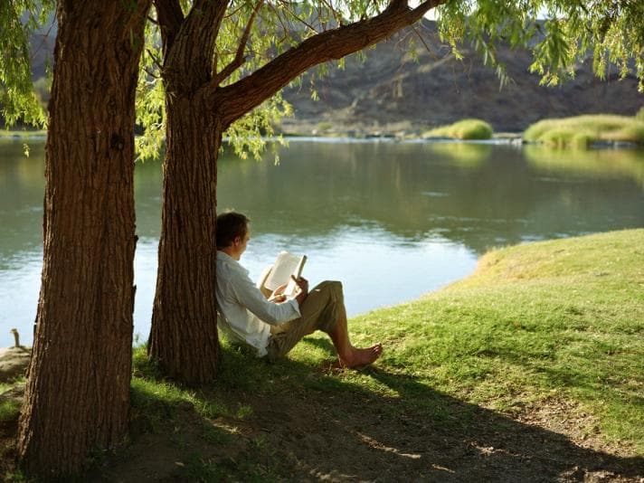 Man reading book under tree by river